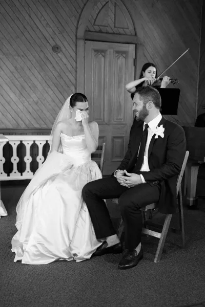 Ceremony moment as a bride wiping tears with a handkerchief while seated beside her groom, set against a wood-paneled wall and arched door.