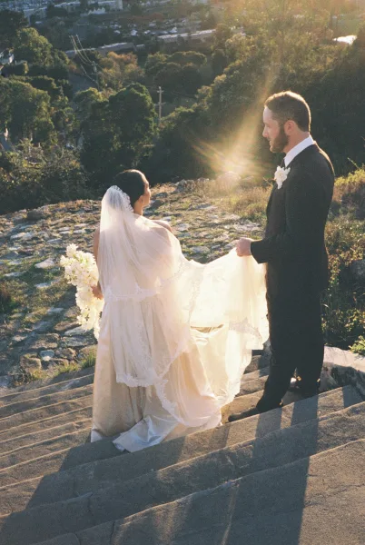 Couple portrait of a bride and groom walking up stone steps, cityscape beyond, as he guides her flowing dress train beneath an open sky.