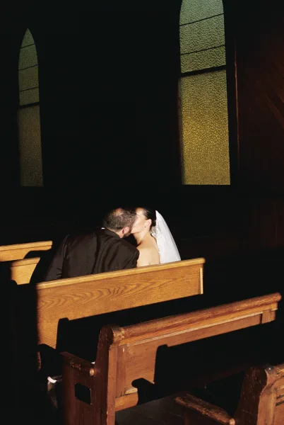 Wedding kiss portrait of a couple kissing in pews, the bride’s veil catching stained glass light in a dark, wood‑paneled church.