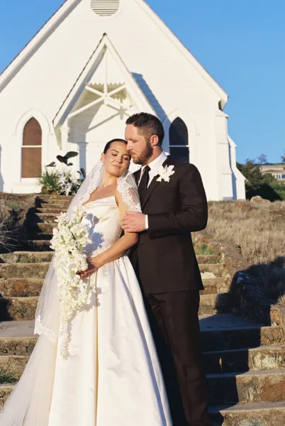 Couple portrait as a church wedding portrait on white chapel stone steps, bride leaning on groom with a lace veil, dry grass hillside.