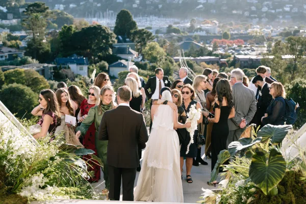 Wedding cocktail hour with guests mingling outdoors, the bridal bouquet glowing on an outdoor terrace above a harbor with boats and hillside homes.