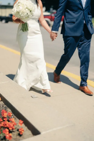 Ceremony recessional as bride and groom walking away holding hands, her white hydrangea bouquet and dress train flowing on a city street sidewalk in daylight