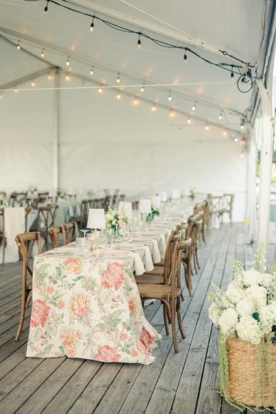 Reception tablescape with floral tablecloth, taper candles, table lamps, and hydrangea baskets under string lights in a white tent
