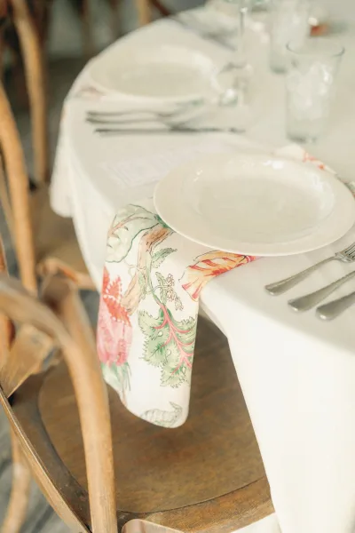 Reception tablescape with wedding place setting of layered white plates, floral napkin accent, forks and clear glasses on white linen near wood chair