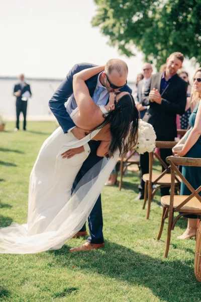 Wedding kiss during a dip kiss wedding as bride in strapless dress holds white bouquet, veil trailing on lawn ceremony by water view