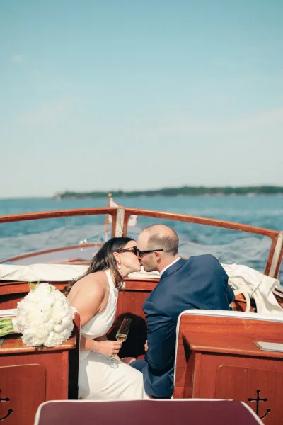 Wedding kiss as the just married kiss unfolds on a wooden boat, bride holding bouquet, both in sunglasses on a bright lake backdrop