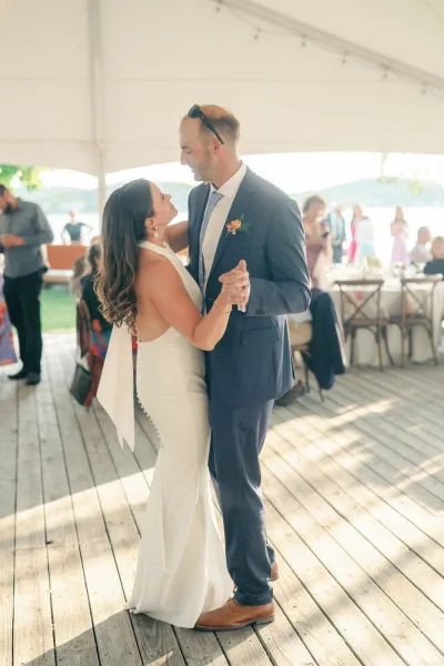 First dance as bride in halter wedding dress and groom in navy suit sway under string lights in a white tent, guests watching