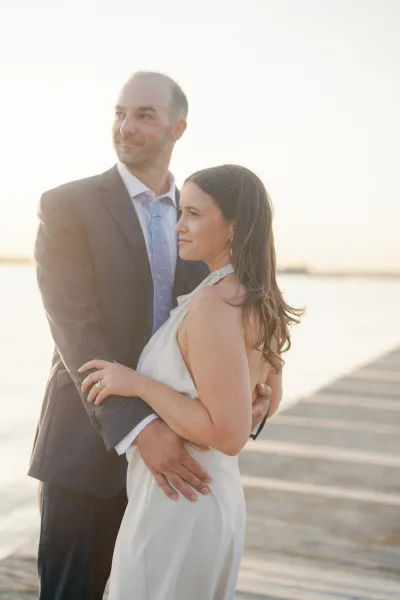 Couple portrait of bride and groom embrace, her halter wedding dress and his suit and necktie glowing in sunset light by the ocean