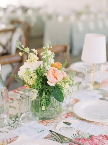 Reception tablescape with wedding floral centerpiece of peach roses, white blooms, and greenery in a clear vase on patterned linen in soft daylight