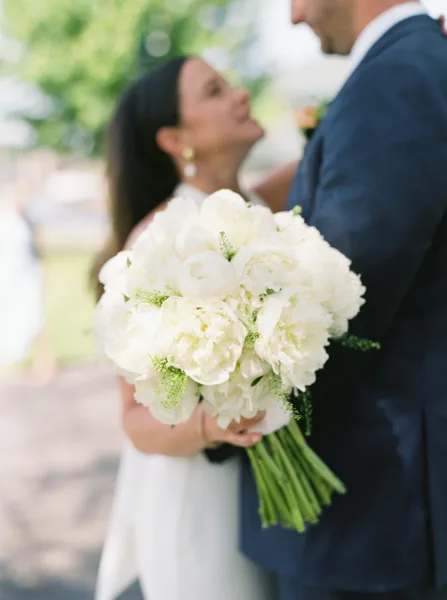 Bridal bouquet of white peonies with greenery accent, held by bride in strapless gown as groom in navy suit embraces outdoors