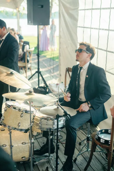 Wedding band drummer in sunglasses playing a drum set under a draped outdoor tent, cymbals and drumsticks mid-beat as guests watch