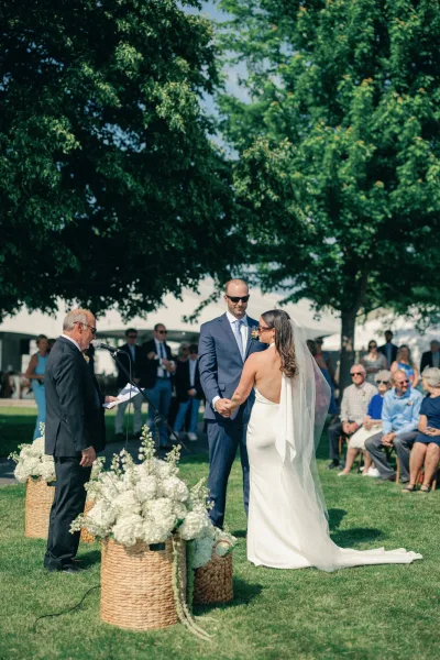 Ceremony moment at an outdoor wedding ceremony as bride and groom hold hands, her long veil trailing on grass under trees by guests
