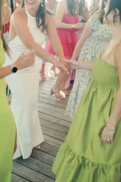 Wedding reception dancing as friends hold hands in a circle, bride in white halter dress with bridesmaids in green on a sunny wood deck