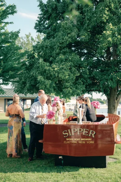 Wedding bar setup in a wooden boat bar with beer taps, pink floral vases and glassware on a lakeside lawn under a large tree