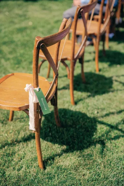 Ceremony seating with outdoor ceremony chairs, wood chairs marked with a reserved tag and ribbon on a sunlit grass lawn with blurred guests
