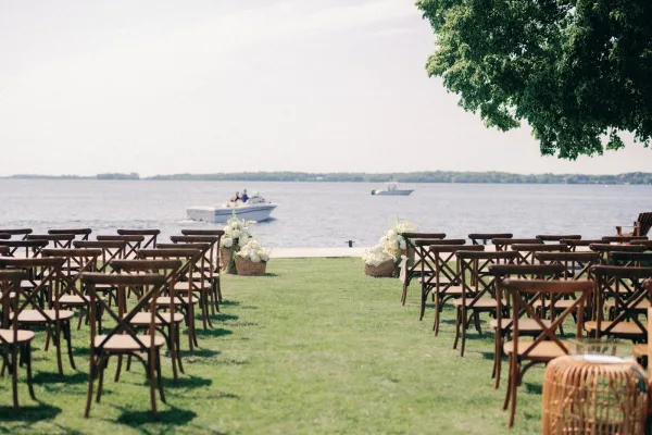 Ceremony setup with outdoor ceremony seating of wood crossback chairs and white aisle florals in baskets by the waterfront lawn with boats beyond