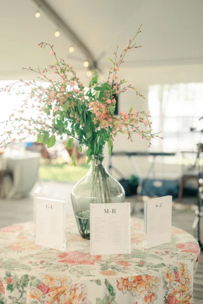 Wedding escort cards on a floral tablecloth, acrylic escort cards arranged alphabetically beside a glass vase with cherry blossoms in a white tent