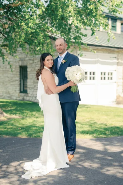 Couple portrait of bride and groom hugging, her long veil and white hydrangea bouquet beside his blue suit in tree shade by a stone building