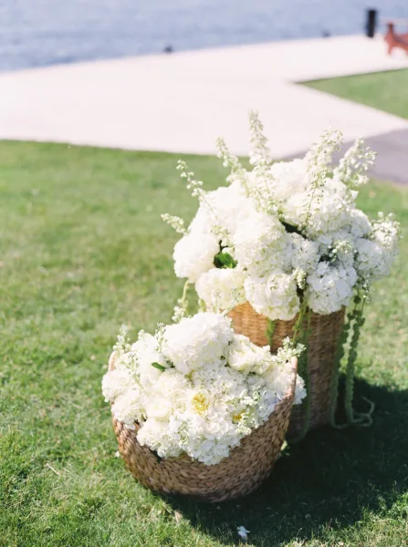 Wedding flower decor with white hydrangeas and roses in wicker baskets lining a paved walkway on a grassy lawn by the water