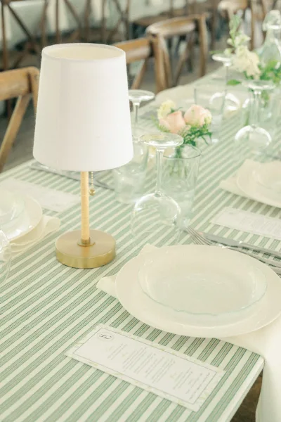 Reception tablescape with wedding table lamp centerpiece on a striped tablecloth, white plates, menu cards, goblets, and pastel roses indoors
