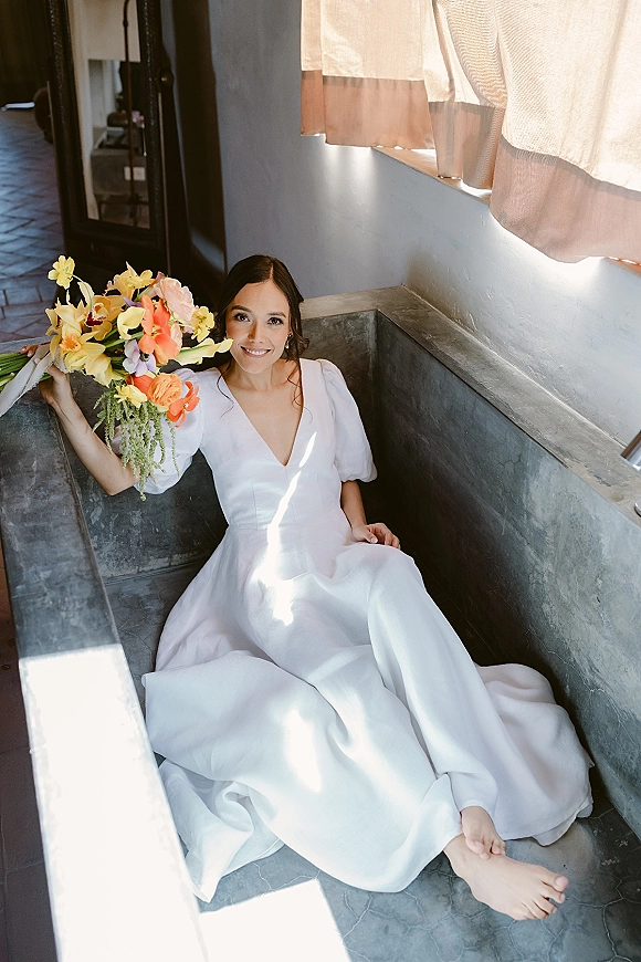 Bridal portrait of a bride holding bouquet of orchids in a metal bathtub, wearing a puff-sleeve gown by sunlit window curtains