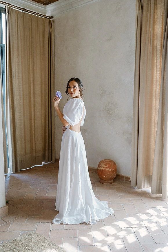 Bridal portrait of a bride in an open back wedding dress with puff sleeves, holding a purple flower by curtains in window light indoors
