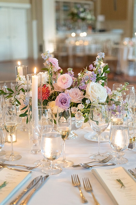 Reception tablescape with wedding table centerpiece of pastel roses and ranunculus, taper candles, glassware and menus in warm indoor room