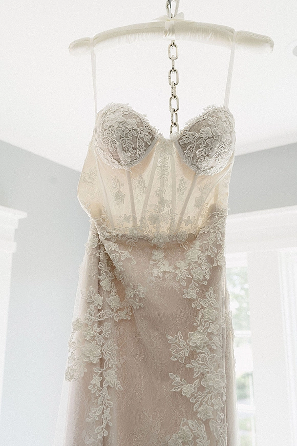 Wedding dress with lace bodice and floral appliqué hanging on a chain hanger, softly lit by window light against a white wall