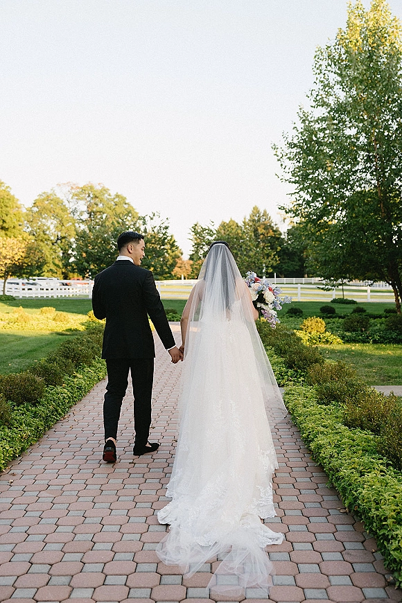 Wedding couple portrait from behind, bride and groom walking away hand in hand, her long veil trailing on a brick garden path