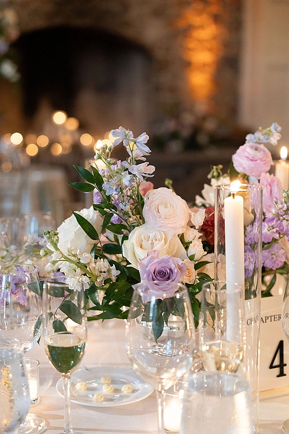 Reception tablescape with wedding floral centerpiece of pastel roses and ranunculus, tall glass taper candles, and blurred guests in warm indoor light