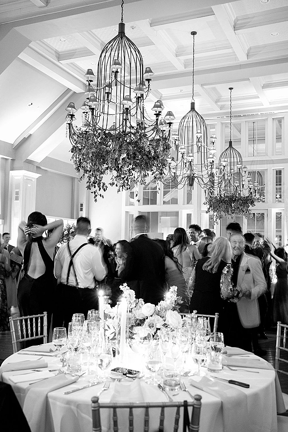 Wedding reception dancing as guests crowd the dance floor beneath chandeliers and hanging greenery in a bright indoor hall with windows