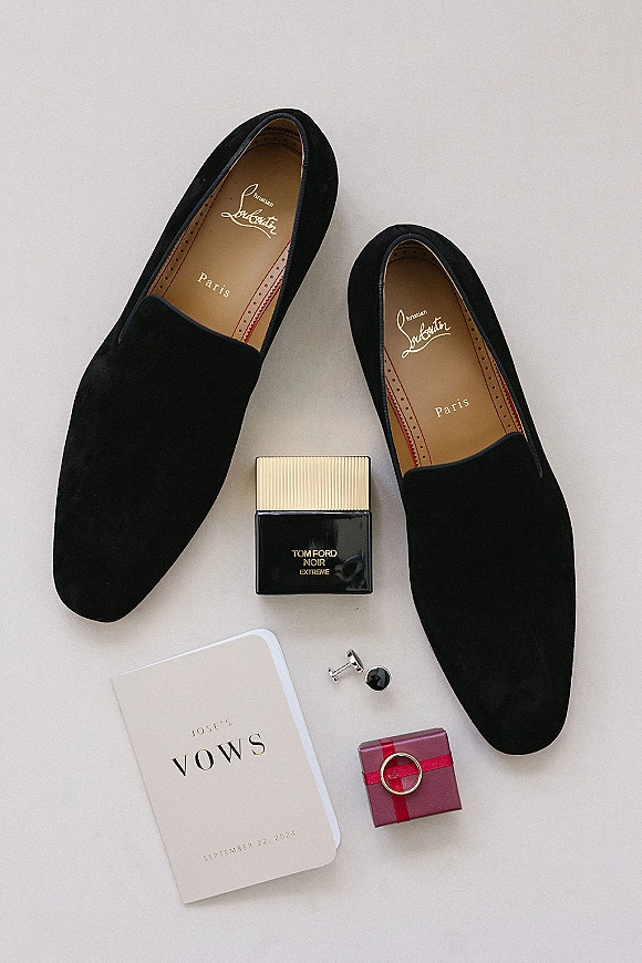 Groom accessories flat lay with black loafers, cologne, cufflinks, vow book, ring box, and wedding band on a light neutral surface