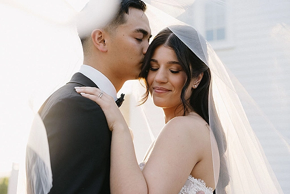 Wedding couple portrait of groom kissing the bride’s forehead as she holds his shoulder, bridal veil flowing on an outdoor terrace with white railing
