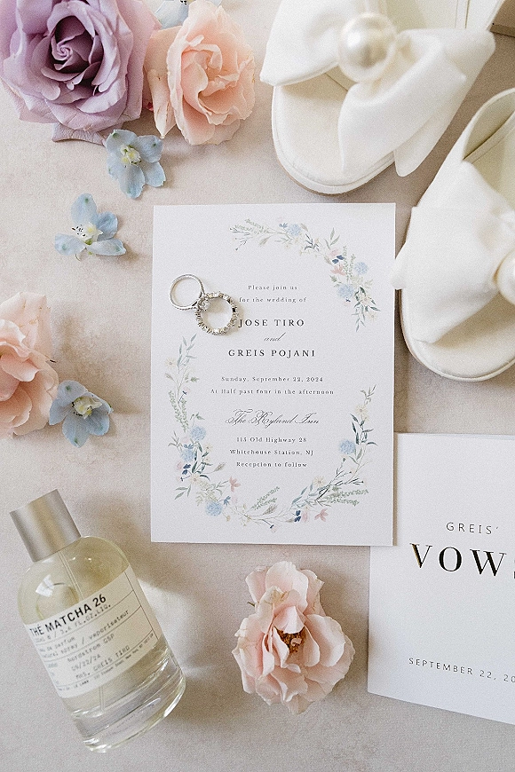 Wedding invitation suite with a floral wedding invitation, vow booklet, rings, bow heels, roses, blue flowers and perfume on neutral tabletop