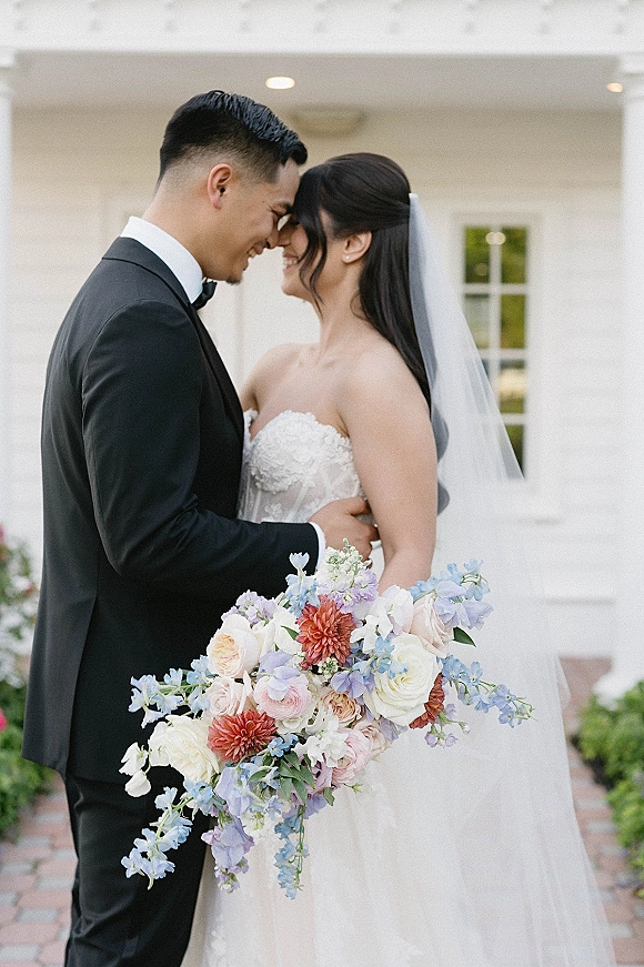 Couple portrait of bride and groom embrace with a forehead touch, her pastel bouquet and long veil against a white wall by a brick walkway