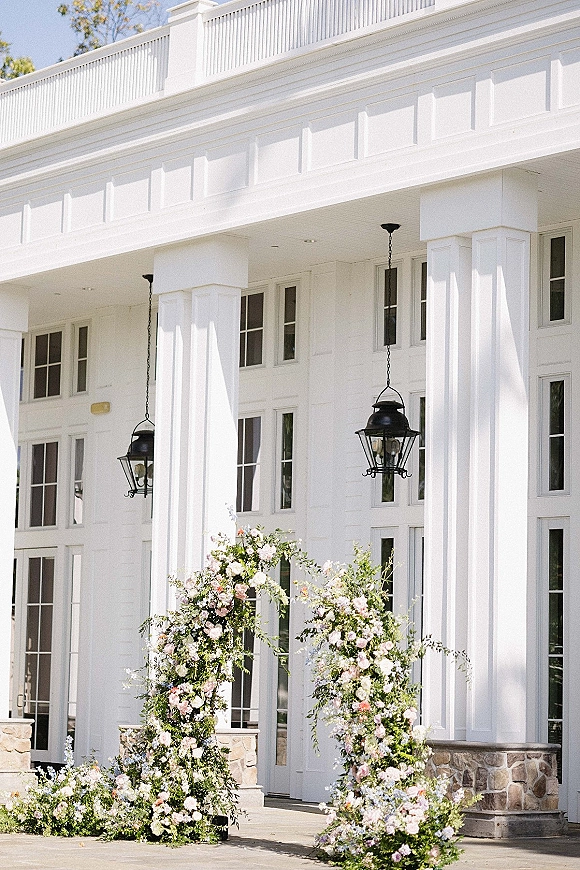 Wedding ceremony arch with asymmetrical floral arch of pastel flowers and greenery, hanging lanterns on a porch with white columns
