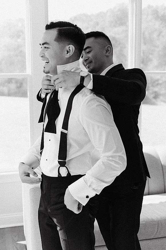 Groom getting ready as groomsmen help groom adjust suspenders, wearing a black tuxedo jacket by large windows with sheer curtains