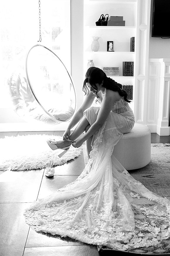 Bride getting ready slips on heels beside her lace strapless gown and long train, lit by bright window light in a cozy living room