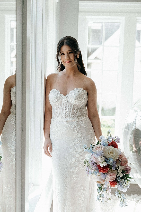 Bridal portrait of a bride in a strapless lace wedding dress holding a rose bouquet with blue blooms by a bright window and mirror