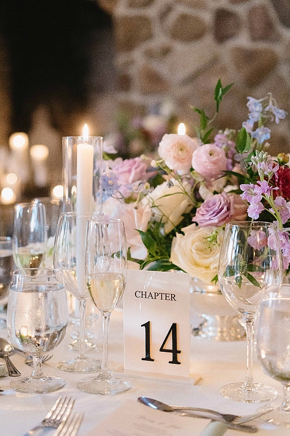 Reception tablescape with wedding table centerpiece of roses and greenery, taper candles in glass holders, and place settings against a stone wall