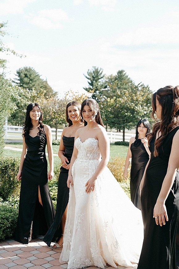 Bride with bridesmaids in black dresses posing on a brick patio, highlighting her strapless lace wedding dress in a garden setting
