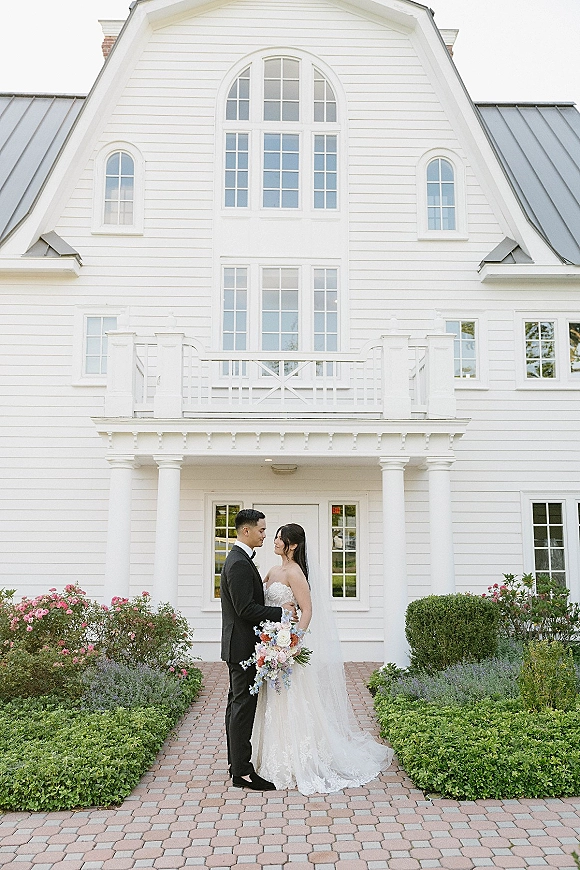 Couple portrait of bride and groom holding a pastel bouquet, bride in strapless lace dress and veil, on a brick path by a white house porch