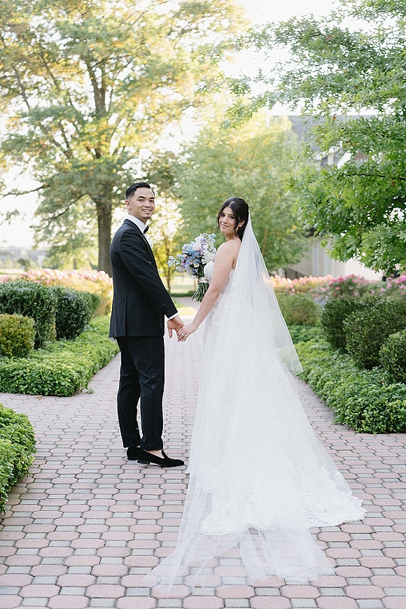Couple portrait of bride and groom holding hands, walking down a brick garden path, her long veil and blue bouquet trailing behind