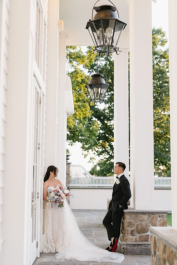 Couple portrait of bride and groom portrait, bride holding bouquet in strapless lace gown as groom in tux poses on a white column porch