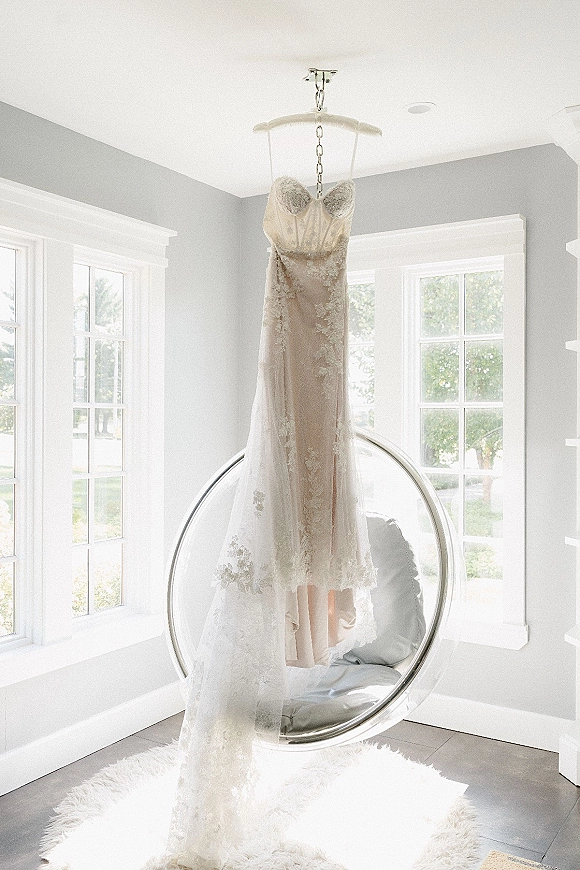 Wedding dress, lace wedding dress hanging from a padded hanger by large windows, with an acrylic bubble chair in a bright room