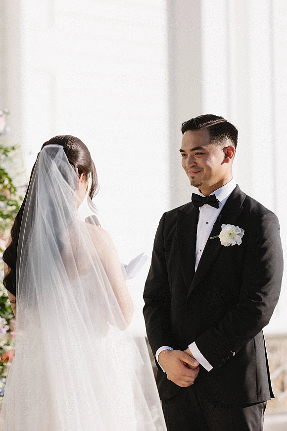 Wedding vows as bride reads into a microphone, cathedral veil flowing, groom in tux with boutonniere before white columns and florals