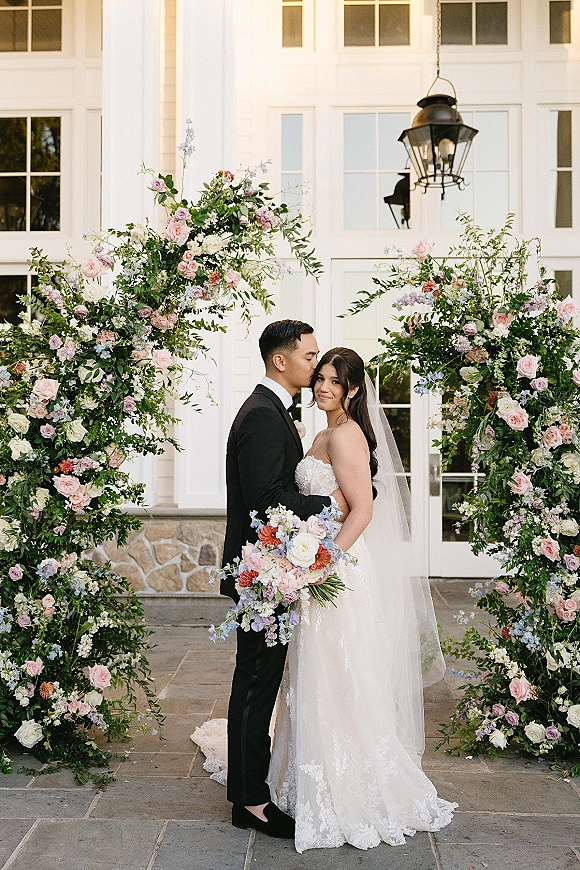 Couple portrait of bride and groom embrace in a forehead kiss, framed by a wedding bouquet and floral arch at a white doorway backdrop