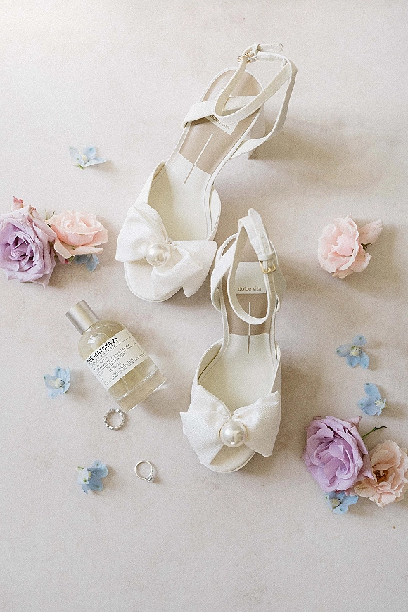 Bridal shoes in a flat lay of white wedding heels with pearl bow details beside rings, perfume, roses, and small blue flowers on neutral surface