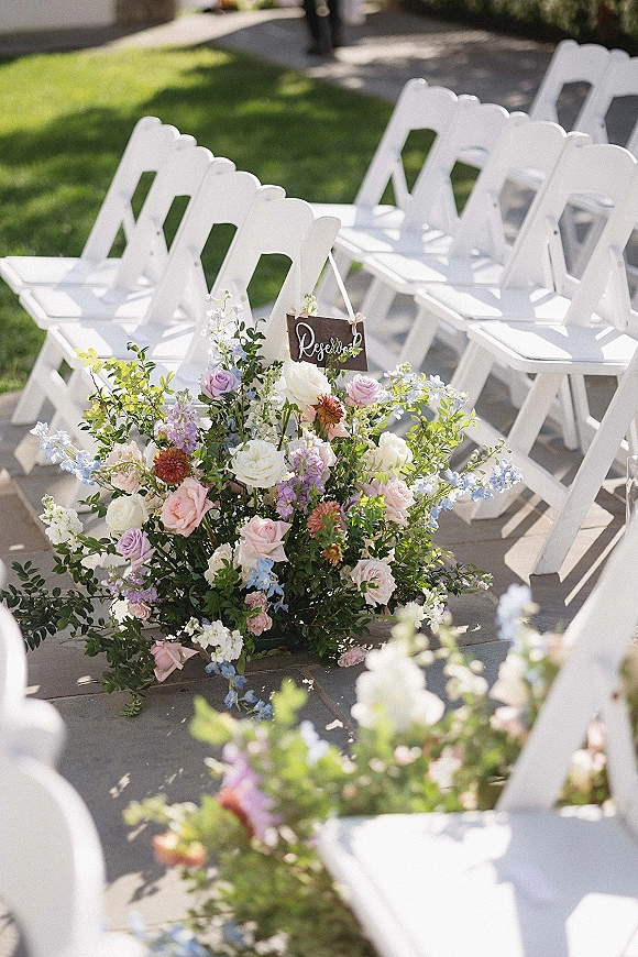Ceremony aisle decor with outdoor ceremony aisle of white folding chairs and pastel rose florals lining a sunlit patio walkway with reserved sign