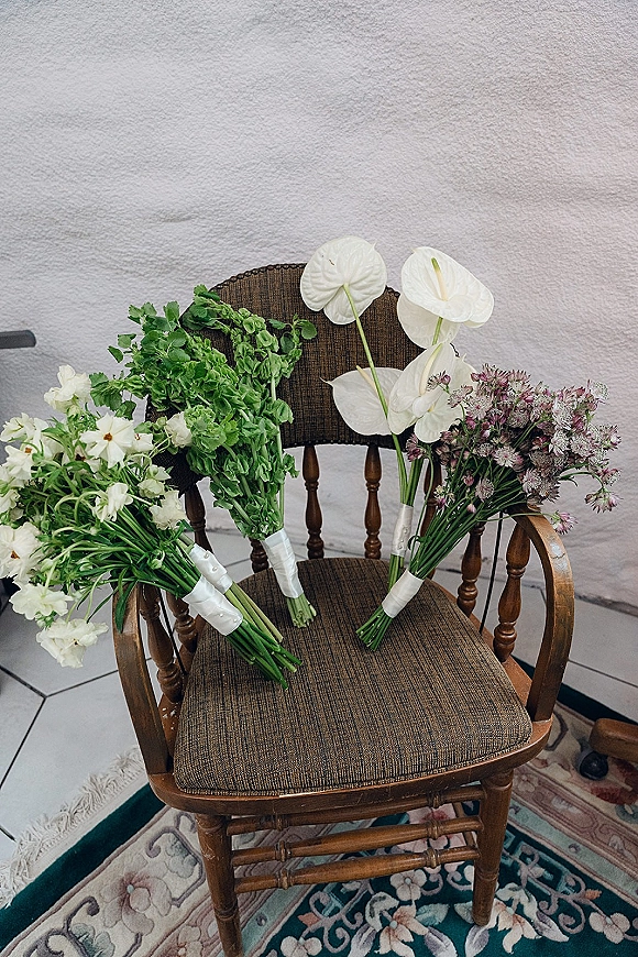 Wedding bouquets with hand tied wedding bouquet stems wrapped in white ribbon, featuring anthurium, set on a wooden chair by a textured wall